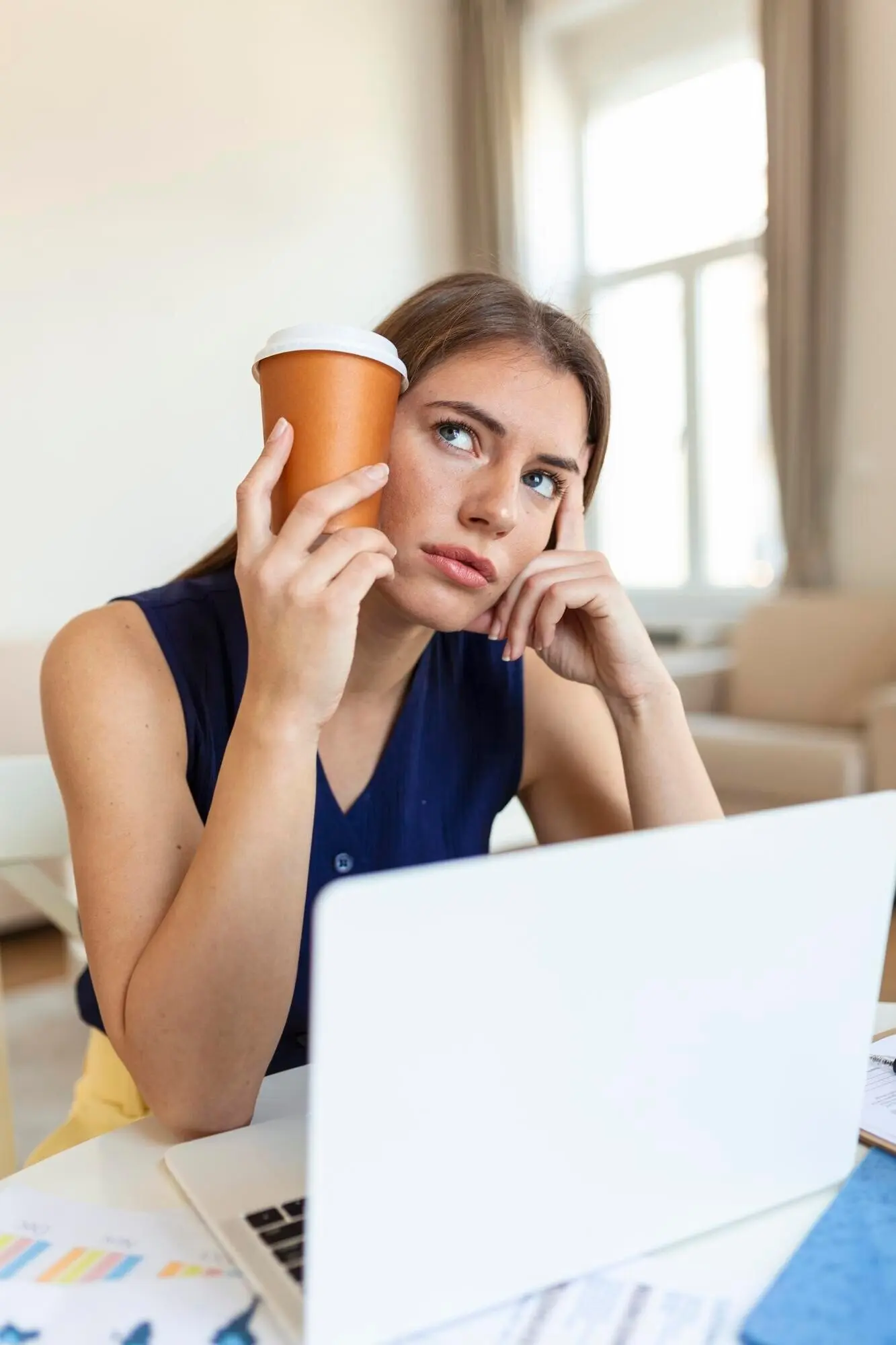 Jeune femme fatiguée regardant ailleurs et tenant une tasse de café près de sa tête dans un bureau, avec un premier plan flou.