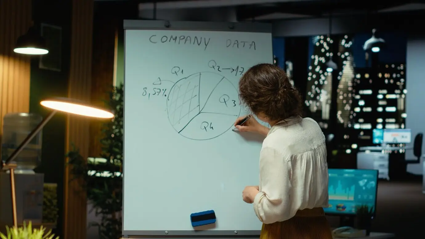 Femme griffonnant des prévisions sur un tableau blanc et vérifiant des analyses sur un ordinateur portable, employée évaluant les finances.